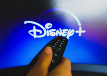 In this photo illustration, a hand holding a TV remote control in front of the Disney Plus logo on a TV screen