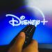 In this photo illustration, a hand holding a TV remote control in front of the Disney Plus logo on a TV screen