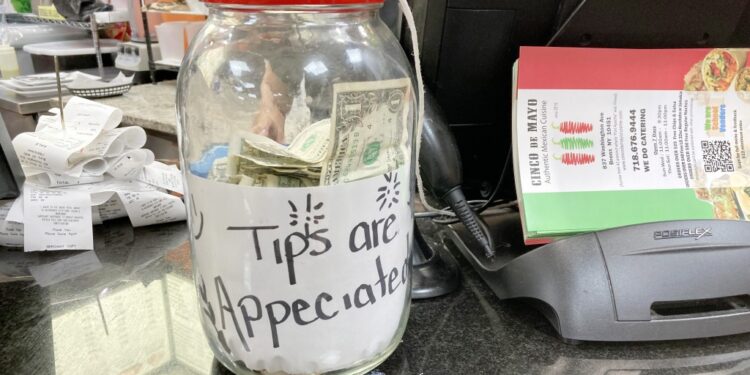 A clear jar filled with money and a label that appears to be made of paper sitting on a table in a restaurant in the Bronx, NY.
