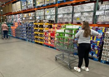 Shoppers browsing and considering purchases in a Costco warehouse, with one woman standing next to a cart full of boxes
