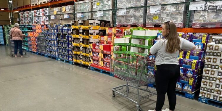 Shoppers browsing and considering purchases in a Costco warehouse, with one woman standing next to a cart full of boxes