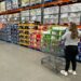 Shoppers browsing and considering purchases in a Costco warehouse, with one woman standing next to a cart full of boxes