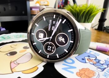 Close up of the OnePlus Watch 3 in emerald green. It sits on top of a black notebook with colorful cartoon stickers. You can also see a plant and pens in the background.