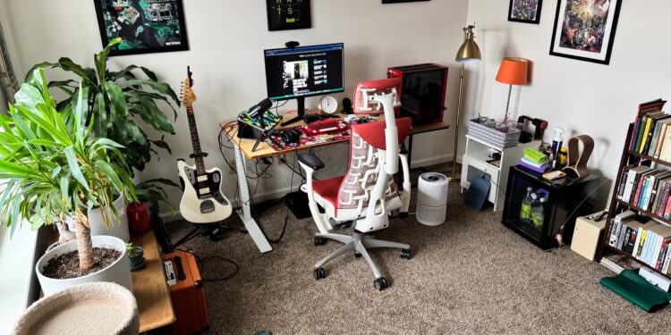 Room in home with desk, red desk chair, plants on the left, bookcase on right, posters on wall.
