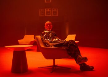 A man in a metallic coat and black trousers sitting in a chair next to a table in a room bathed in red light.