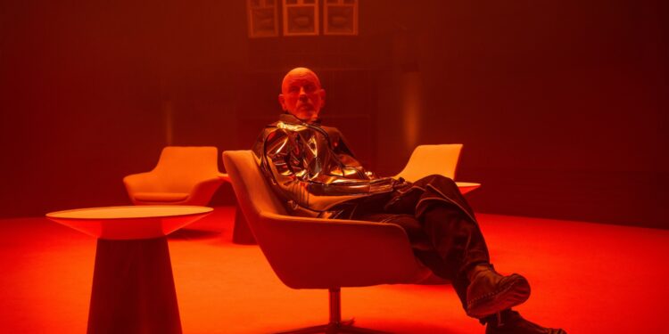 A man in a metallic coat and black trousers sitting in a chair next to a table in a room bathed in red light.