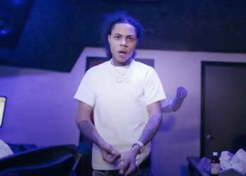 A photo of drill rapper Kay Flock wearing a white t-shirt, silver necklace and with a large watch