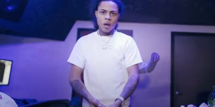 A photo of drill rapper Kay Flock wearing a white t-shirt, silver necklace and with a large watch