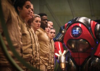 A group of people wearing all-tan outfits and standing in a line as a hulking robot leers at them .