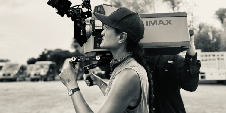 A woman in a white tank top holding an IMAX camera on her shoulder.