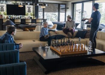 Three men sitting on a couch in a luxurious living room while a fourth man stands in front of them.