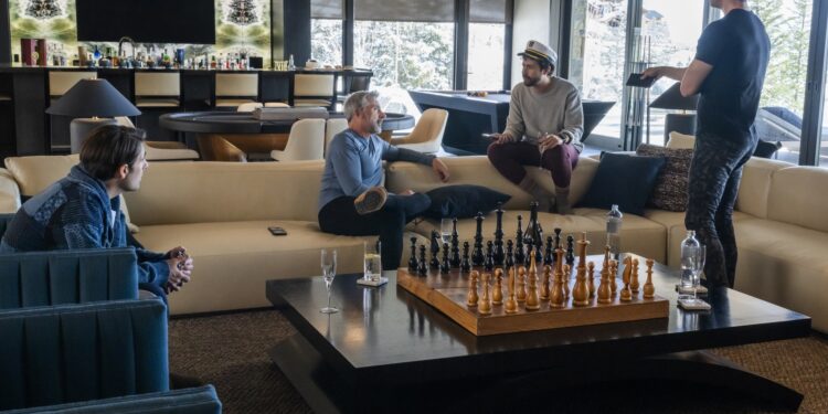 Three men sitting on a couch in a luxurious living room while a fourth man stands in front of them.