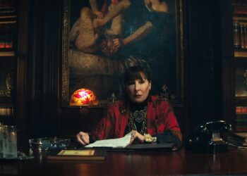 A woman wearing a black shirt and a red sweater sitting at a massive desk in an office lined with leather-bound books.