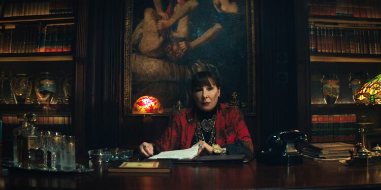 A woman wearing a black shirt and a red sweater sitting at a massive desk in an office lined with leather-bound books.