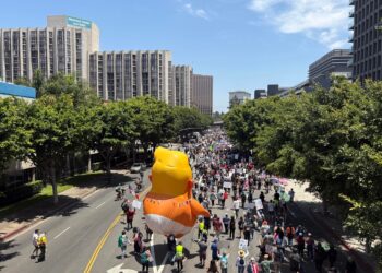A parade balloon depicting Donald Trump as a baby wearing a diaper floats above a crowd of people marching down a street.