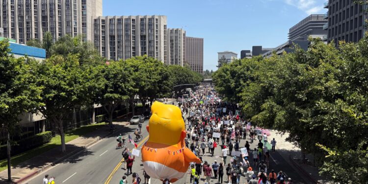 A parade balloon depicting Donald Trump as a baby wearing a diaper floats above a crowd of people marching down a street.