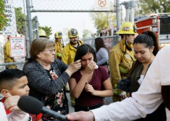 A crying girl comforted by a woman, with firefighters in the background.