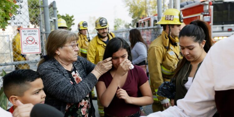A crying girl comforted by a woman, with firefighters in the background.