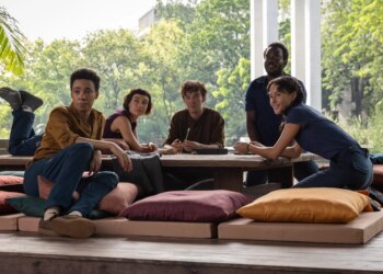 Two women and three men all sitting together at a table that’s close to the ground and surrounded by pillows to sit on. Behind the group is a lush jungle that can be seen through a large glass window.