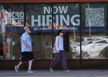 Two people walk past a store window advertising job openings.