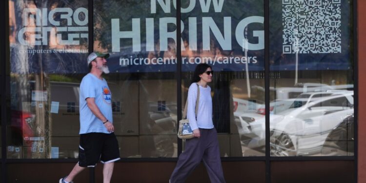 Two people walk past a store window advertising job openings.