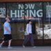 Two people walk past a store window advertising job openings.