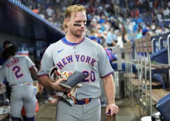 Pete Alonso in the dugout after a loss, wearing a gray Mets uniform, holding a notebook, gloves, and a bat.