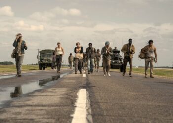 A group of 10 young men walking along a desolate highway in the countryside. Behind the men are two military vehicles being driven by soldiers with guns.