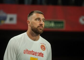 Travis Kelce wearing a white shirt with a Kansas City Chiefs emblem.