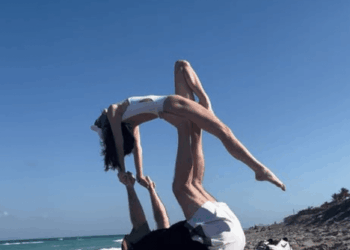 A man on his back on a beach towel supporting a woman in an acrobatic pose on his feet.