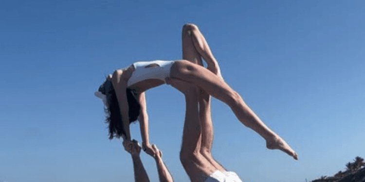 A man on his back on a beach towel supporting a woman in an acrobatic pose on his feet.