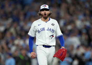 Blue Jays baseball player with long hair and beard wearing a white jersey, blue undershirt, and a World Series patch on his sleeve, holding a red glove and looking forward with his mouth open.