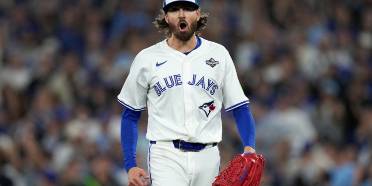 Blue Jays baseball player with long hair and beard wearing a white jersey, blue undershirt, and a World Series patch on his sleeve, holding a red glove and looking forward with his mouth open.