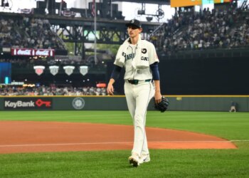 A baseball player in a white uniform with "Mariners" written on it and a dark blue cap walks across the field, holding a glove.