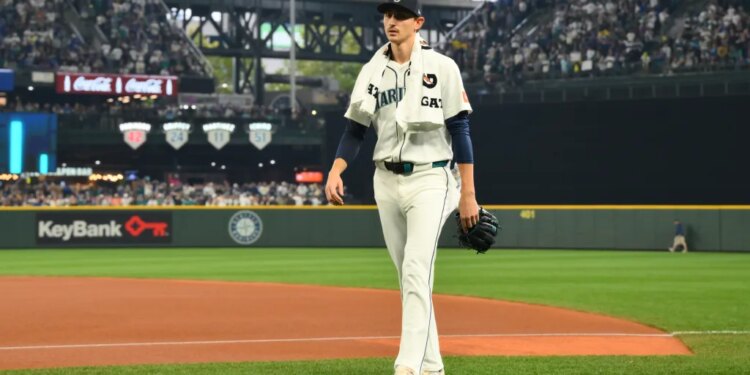 A baseball player in a white uniform with "Mariners" written on it and a dark blue cap walks across the field, holding a glove.