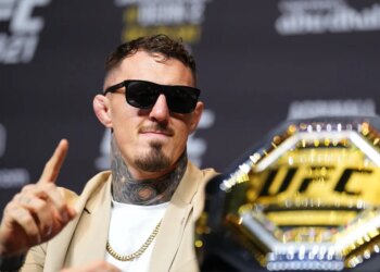 Tom Aspinall wearing sunglasses and a gold chain, pointing his finger with a UFC belt in the foreground.
