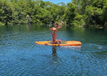 Brie Larson kneeling on an orange paddleboard on a body of water with lush green trees in the background.