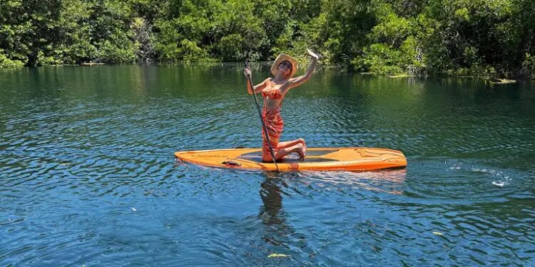 Brie Larson kneeling on an orange paddleboard on a body of water with lush green trees in the background.