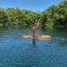 Brie Larson kneeling on an orange paddleboard on a body of water with lush green trees in the background.