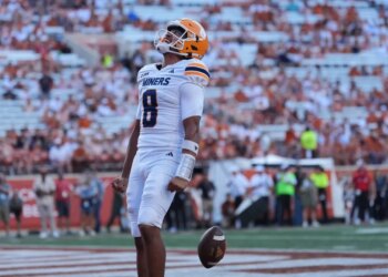 A UTEP football player in a white uniform with "Miners" and number 8, wearing an orange helmet, looks up with a football floating near his waist.
