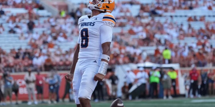 A UTEP football player in a white uniform with "Miners" and number 8, wearing an orange helmet, looks up with a football floating near his waist.