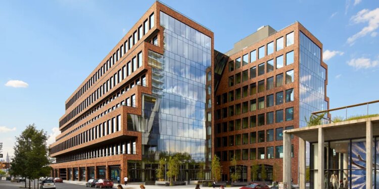 A large brick and glass building with several people walking on the sidewalk and cars parked along the street.