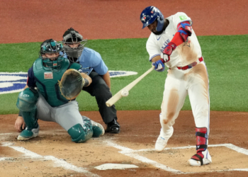 A Toronto Blue Jays batter swings at a baseball, while a Seattle Mariners catcher and a home plate umpire watch.