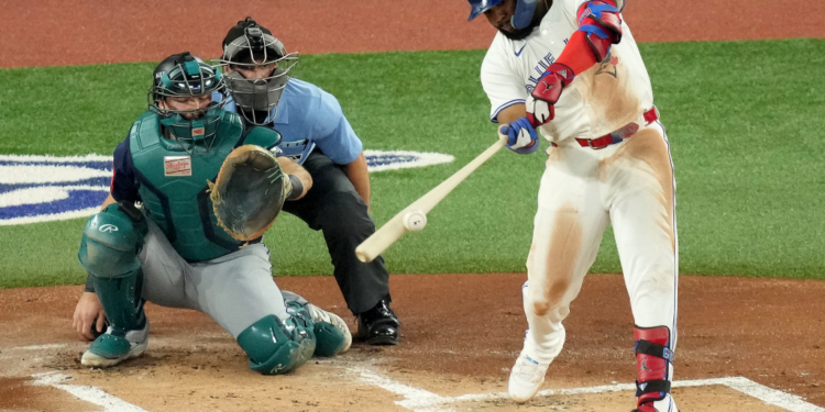 A Toronto Blue Jays batter swings at a baseball, while a Seattle Mariners catcher and a home plate umpire watch.