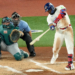 A Toronto Blue Jays batter swings at a baseball, while a Seattle Mariners catcher and a home plate umpire watch.