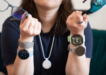 Close up of Senior reviewer Victoria Song holding two pairs of smart glasses, wearing four wrist-based wearables, a smart ring, and the Friend AI necklace.