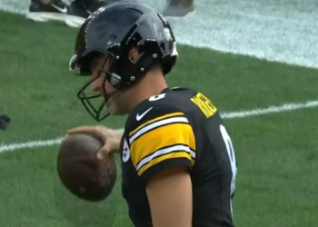 A Pittsburgh Steelers player in a black and yellow jersey holding a football on a green field.