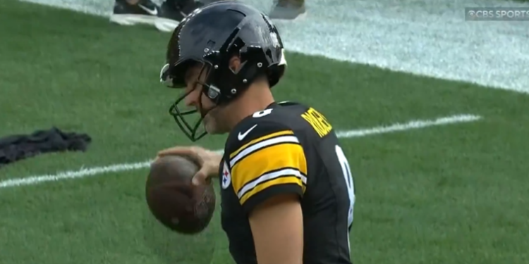 A Pittsburgh Steelers player in a black and yellow jersey holding a football on a green field.
