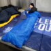 Yankees GM Brian Cashman in a blue sleeping bag on a blue mat that says "Sleep Out" for the Covenant House Sleep Out event.
