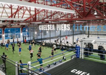 An indoor fitness facility with people exercising on a green turf, a boxing ring, and a basketball court, with "Chelsea Piers Fitness" written overhead.